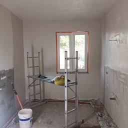 Remont4You - Pokój w trakcie prac wykończeniowych: ściany po szpachlowaniu, rusztowanie, wiadro farby, instalacja elektryczna. Widok na okno z pomarańczową ramą.