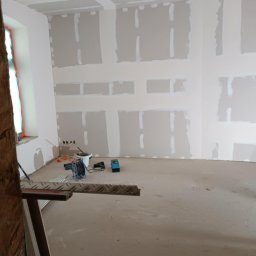 Remont4You - Wnętrze w trakcie prac: ściany i sufit z płyt kartonowo-gipsowych, częściowo wyszpachlowane. Widoczne narzędzia i drabina, okno z lewej strony.