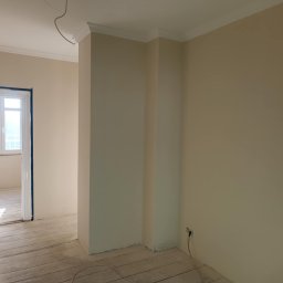 Remont4You - Jasne wnętrze po malowaniu ścian na kremowo. Widoczne listwy przysufitowe, drewniana podłoga i okno z widokiem na zieleń. Częściowo wykończone pomieszczenie.