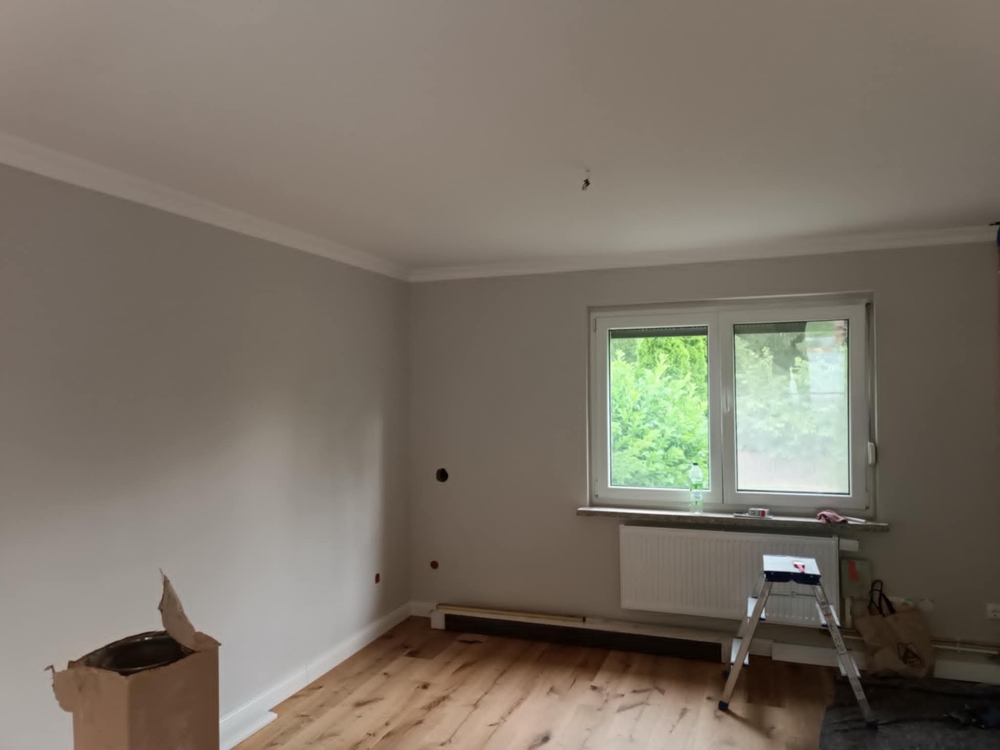 Pokój po malowaniu ścian na szaro z białą listwą sufitową i drewnianą podłogą. Widoczne drabina malarska i puszka z farbą. Okno z widokiem na zieleń.