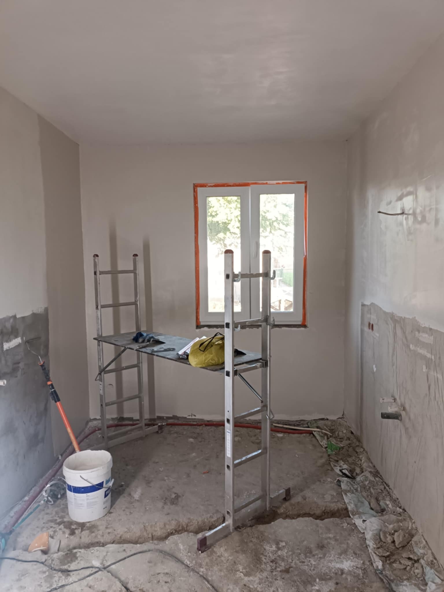 Pokój w trakcie prac wykończeniowych: ściany po szpachlowaniu, rusztowanie, wiadro farby, instalacja elektryczna. Widok na okno z pomarańczową ramą.
