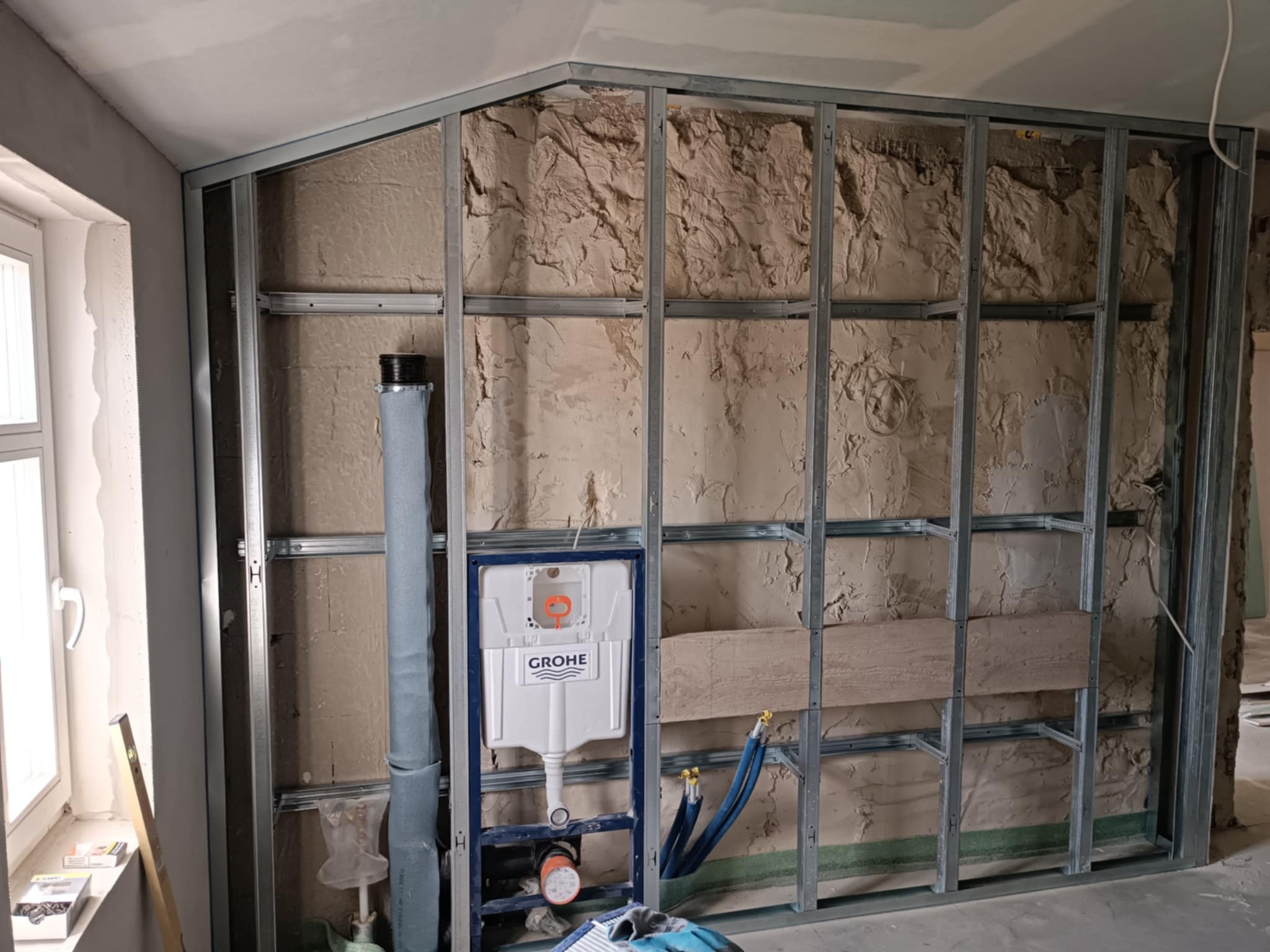 Stelaż metalowy z zabudową GK w łazience, widoczne rury i zbiornik WC Grohe. Ściana z surowym tynkiem. Etap prac wykończeniowych przed położeniem płytek.