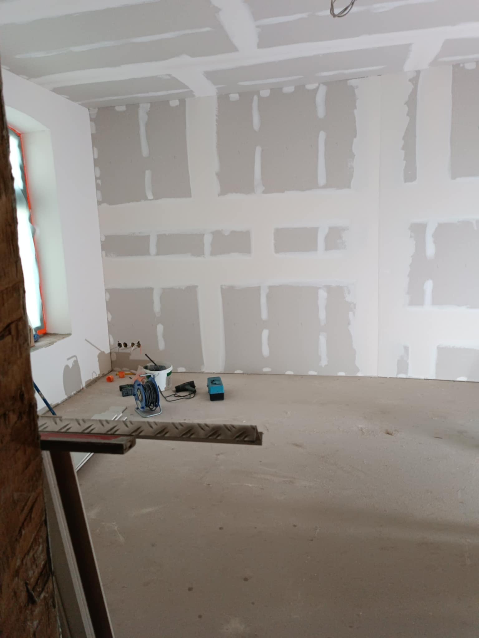Wnętrze w trakcie prac: ściany i sufit z płyt kartonowo-gipsowych, częściowo wyszpachlowane. Widoczne narzędzia i drabina, okno z lewej strony.