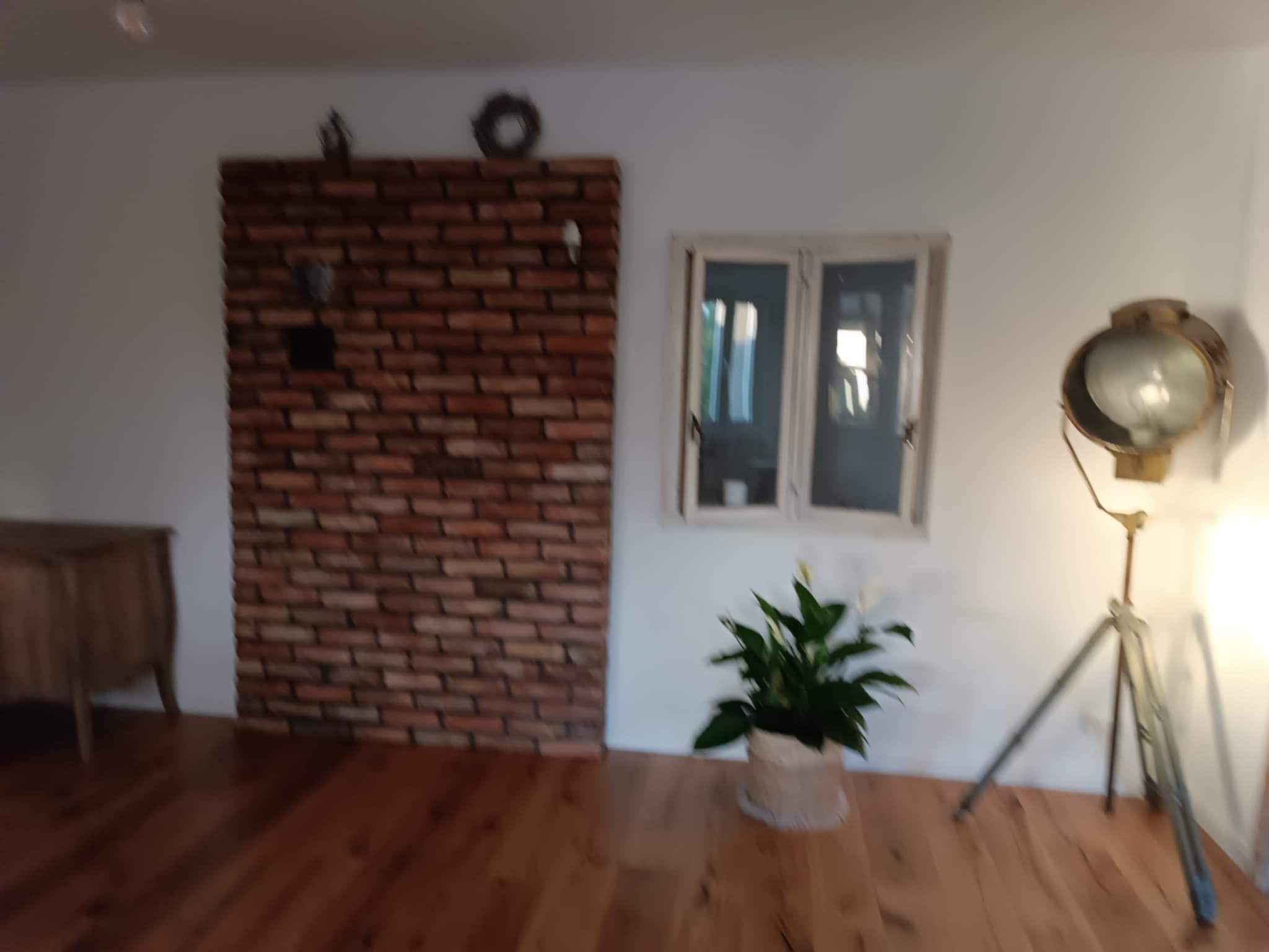 Ceglana ściana z dekoracją, okno z rośliną i lampa na trójnogu tworzą przytulny, odświeżony kącik w jasnym wnętrzu z parkietem.