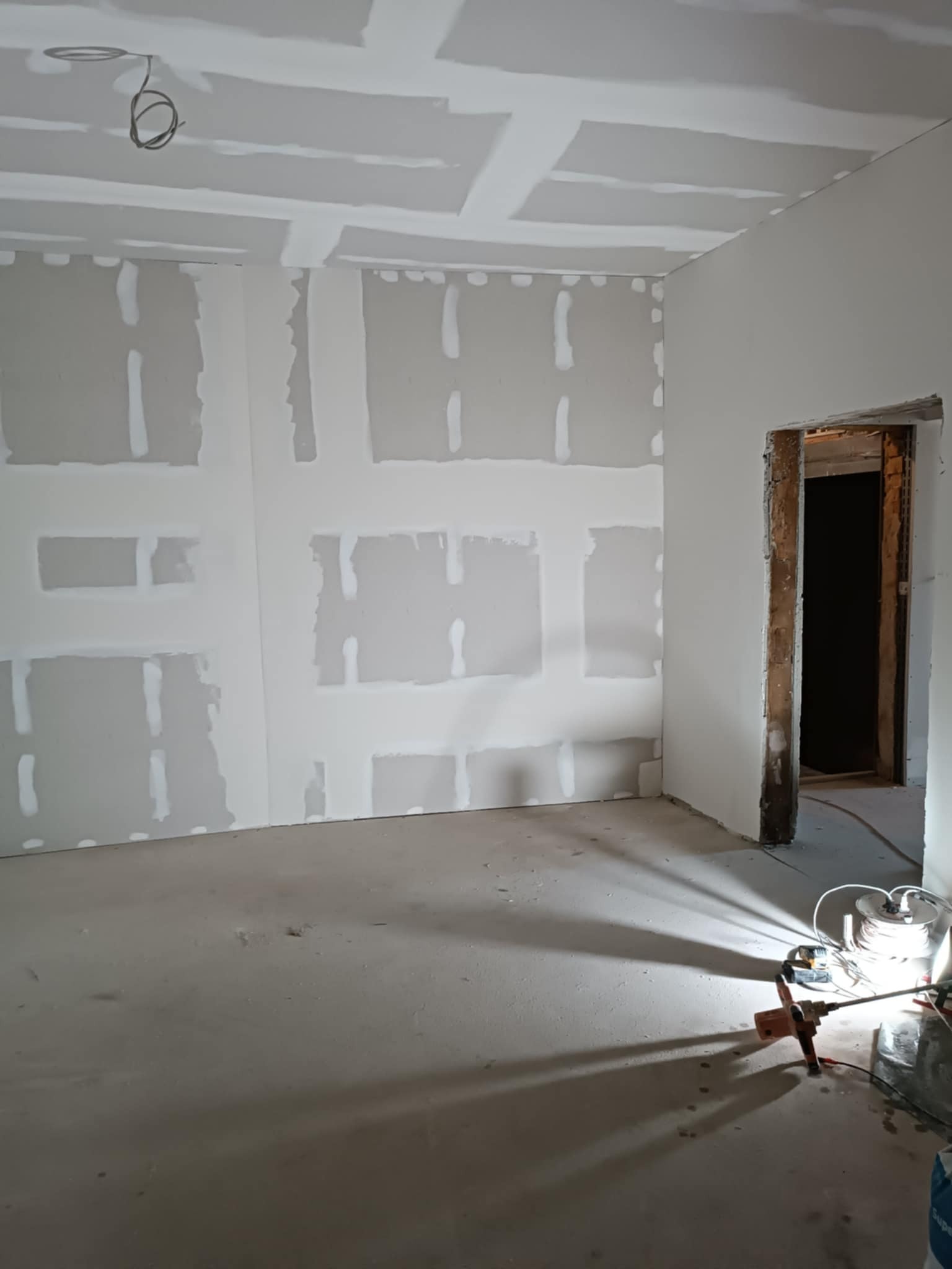 Pokój w trakcie prac: płyty gipsowo-kartonowe na ścianach i suficie, widoczne łączenia szpachlowane, kabel zwisający z sufitu i lampa budowlana rzucająca cień.
