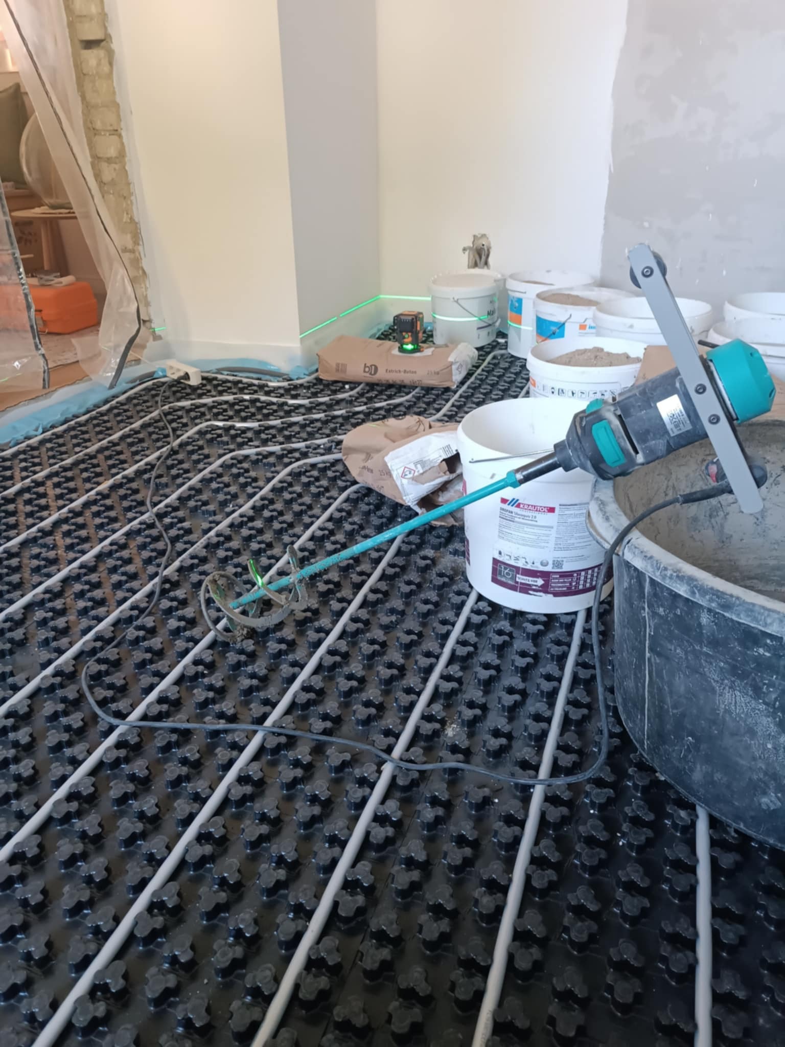 Rozłożona instalacja ogrzewania podłogowego z widocznym laserem poziomującym, workiem zaprawy i mieszarką do betonu. Przygotowanie do wylewki.