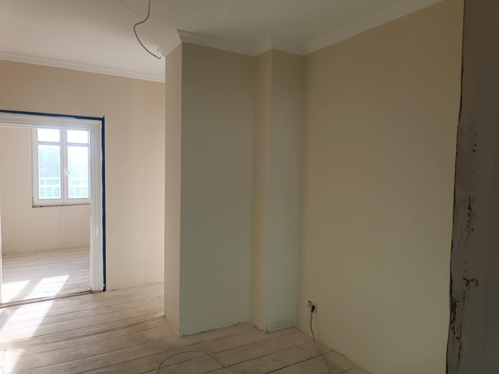 Jasne wnętrze po malowaniu ścian na kremowo. Widoczne listwy przysufitowe, drewniana podłoga i okno z widokiem na zieleń. Częściowo wykończone pomieszczenie.