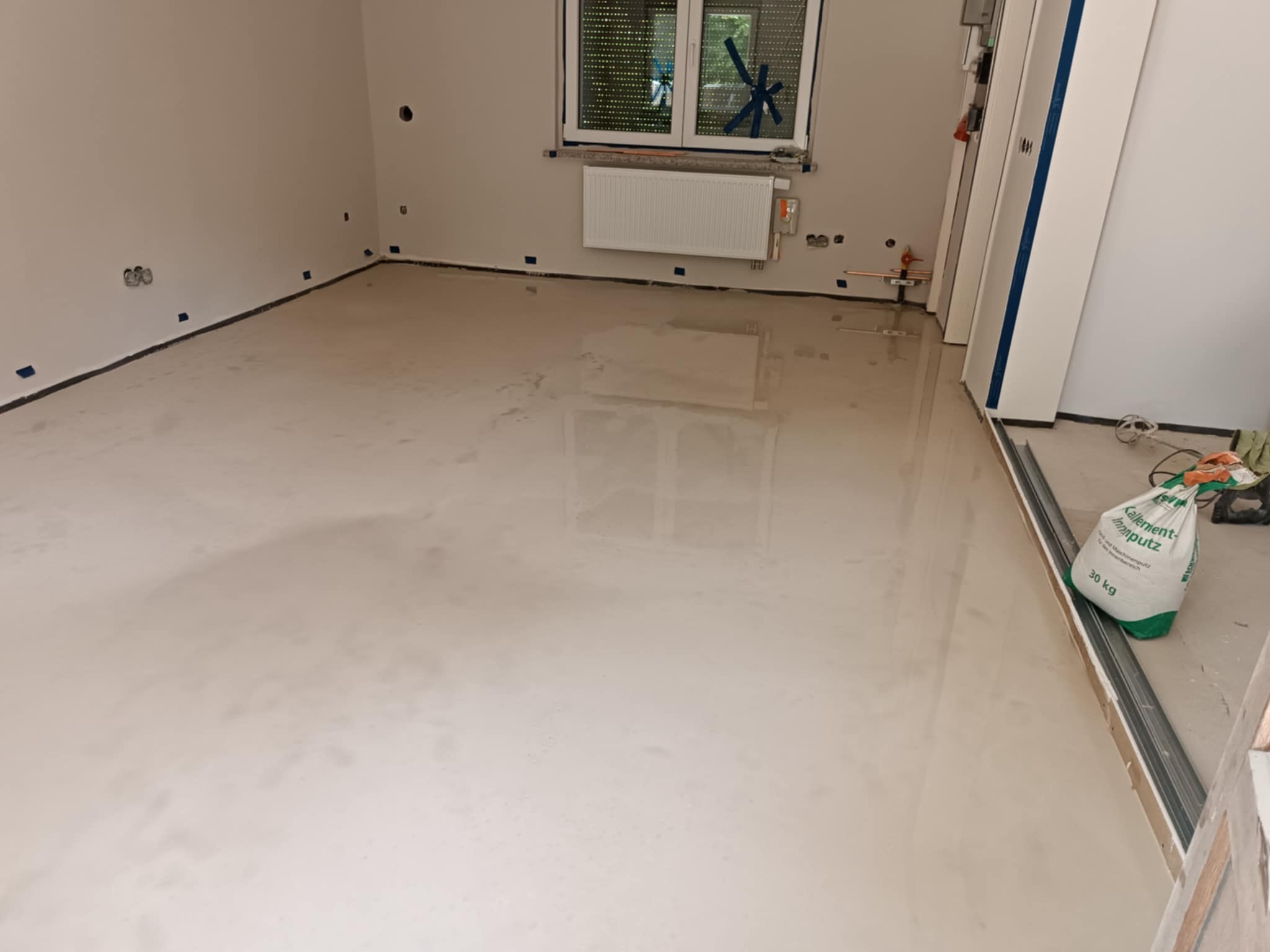 Wylana, gładka posadzka samopoziomująca w jasnym pomieszczeniu z grzejnikiem. W tle okno z roletami. W rogu worki z zaprawą.