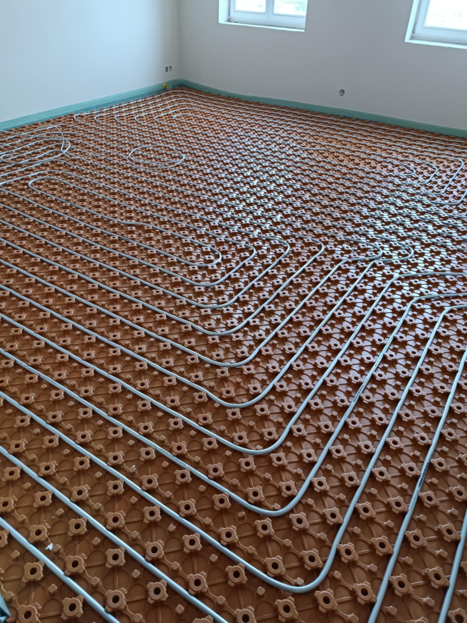 Instalacja ogrzewania podłogowego: rury ułożone na brązowej macie z wypustkami, widok z góry. Etap prac przed wylaniem posadzki.