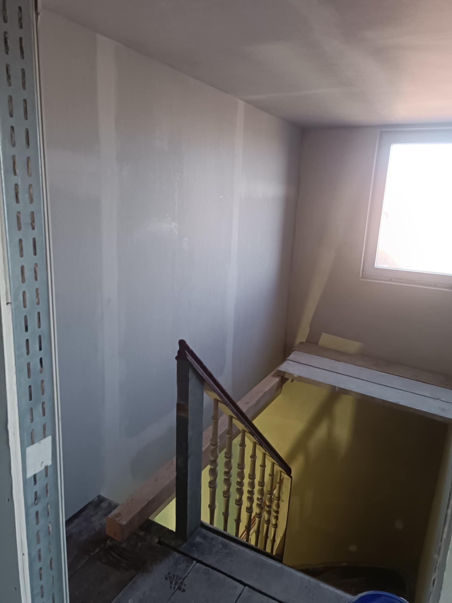 Ściana po szpachlowaniu w jasnym wnętrzu z drewnianymi schodami i metalowym profilem. Widoczne okno w tle. Prace wykończeniowe w trakcie realizacji.