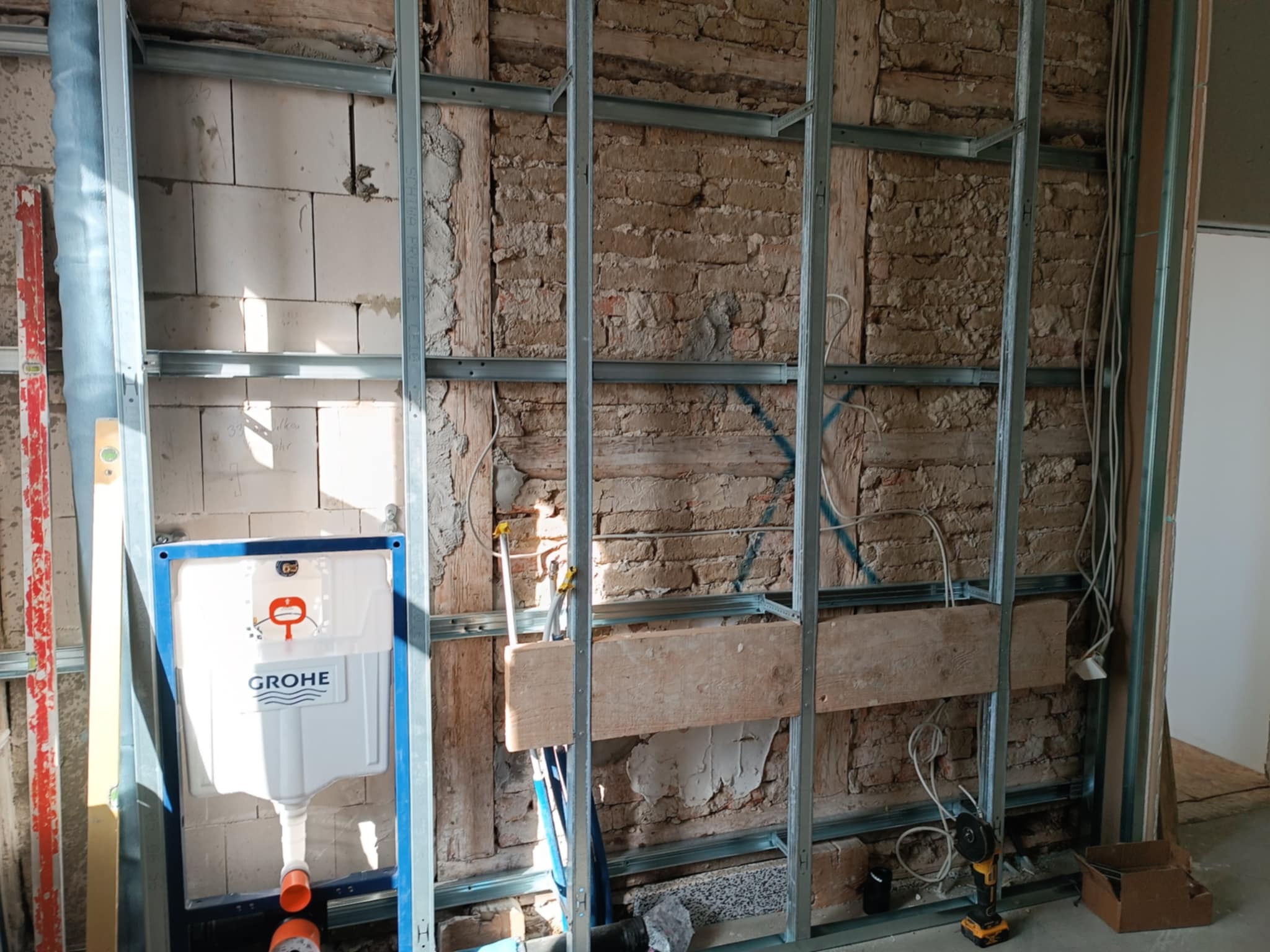 Montaż stelaża podtynkowego Grohe na tle starej ceglanej ściany, widoczne profile metalowe i narzędzia. Prace wykończeniowe w trakcie realizacji.