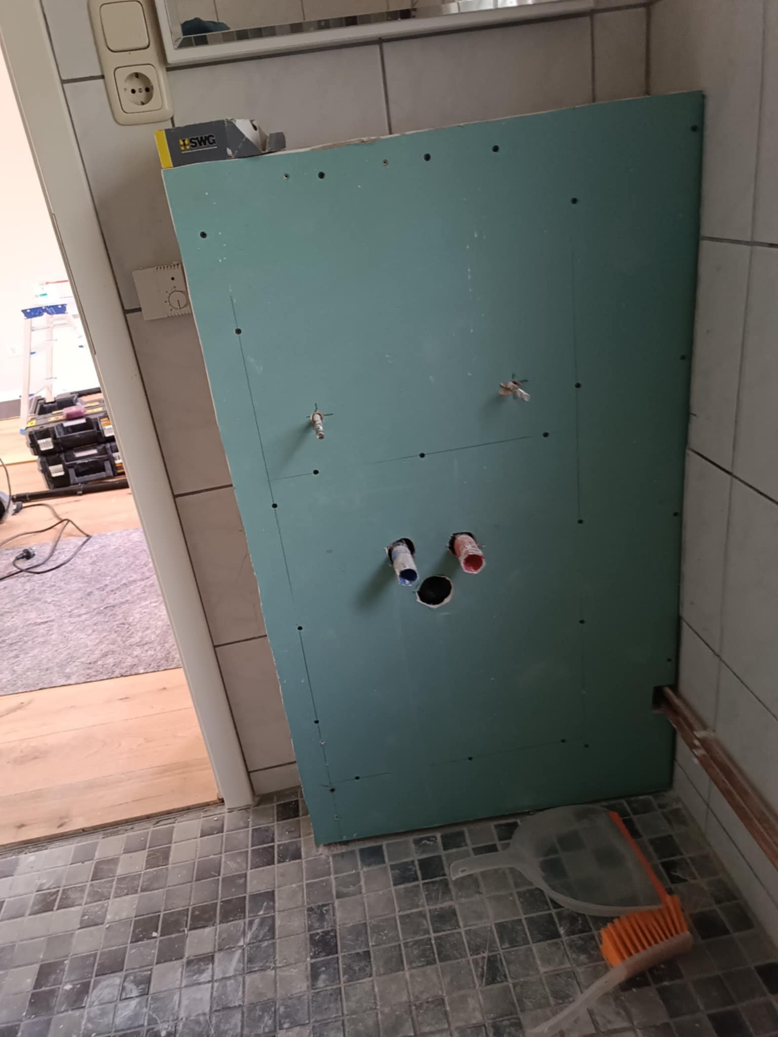 Płyta GK z otworami na instalacje w łazience, widoczne rury i otwory na baterie. Na podłodze płytki i narzędzia. Prace wykończeniowe w toku.