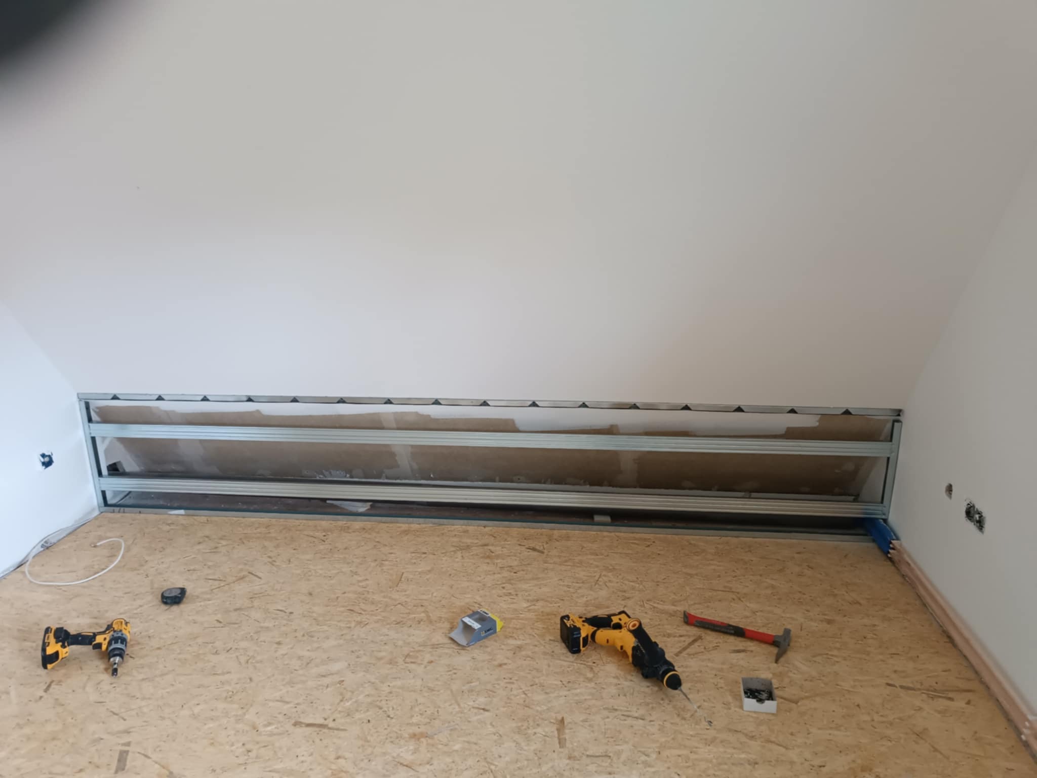Poddasze w trakcie adaptacji: widoczne profile metalowe, płyta OSB na podłodze i narzędzia (wiertarka, młotek). Przygotowanie do zabudowy ścian i sufitu.