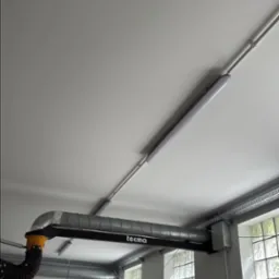 Zamontowany system wentylacyjny Tecma z rurami i oświetleniem LED na suficie, widok z dołu, z drabiną w tle. Widoczne okna z luksferami i skrzynki elektryczne.