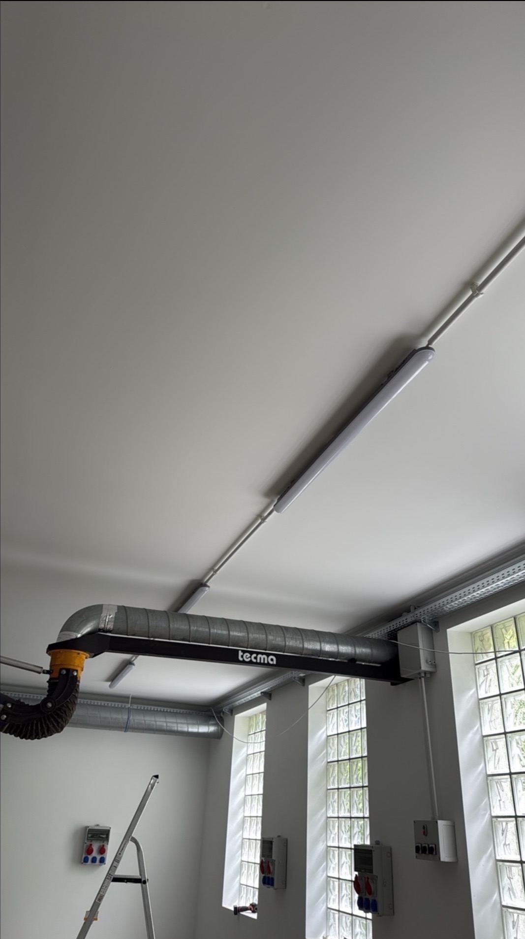 Zamontowany system wentylacyjny Tecma z rurami i oświetleniem LED na suficie, widok z dołu, z drabiną w tle. Widoczne okna z luksferami i skrzynki elektryczne.