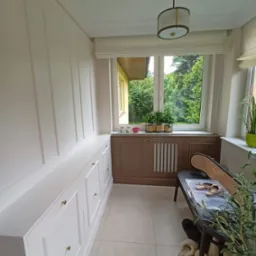 Zabudowa balkonu z szafkami w stylu angielskim, białe fronty z frezami i złotymi uchwytami. Na blacie doniczki z roślinami, w tle okno z widokiem na zieleń.