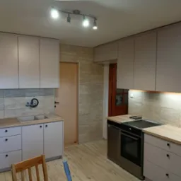 Jasna kuchnia z drewnianym blatem i szarymi frontami. Górne szafki podświetlane, na suficie reflektory. Widoczna płyta grzewcza i piekarnik.