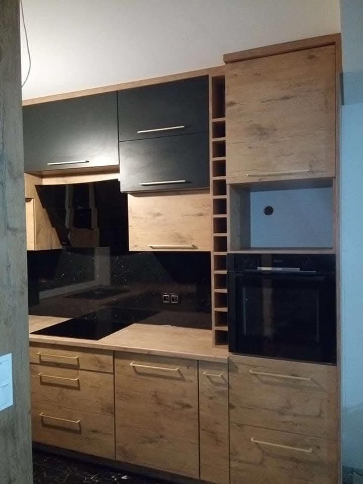 Nowoczesna kuchnia z czarnymi szafkami górnymi i drewnopodobnymi dolnymi, z piekarnikiem w zabudowie i czarnym panelem szklanym nad blatem. Widoczna półka na wino.