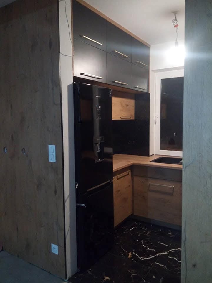 Ciemna kuchnia z drewnianymi akcentami: szafki górne w kolorze grafitu z długimi, złotymi uchwytami, czarna lodówka i marmurowa podłoga w odcieniach czerni i bieli.