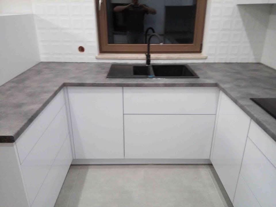 Nowoczesna kuchnia z białymi szafkami i szarym blatem, czarny zlew i bateria. W tle okno z odbiciem osoby. Minimalistyczny design, jasne wnętrze, płytki z fakturą.