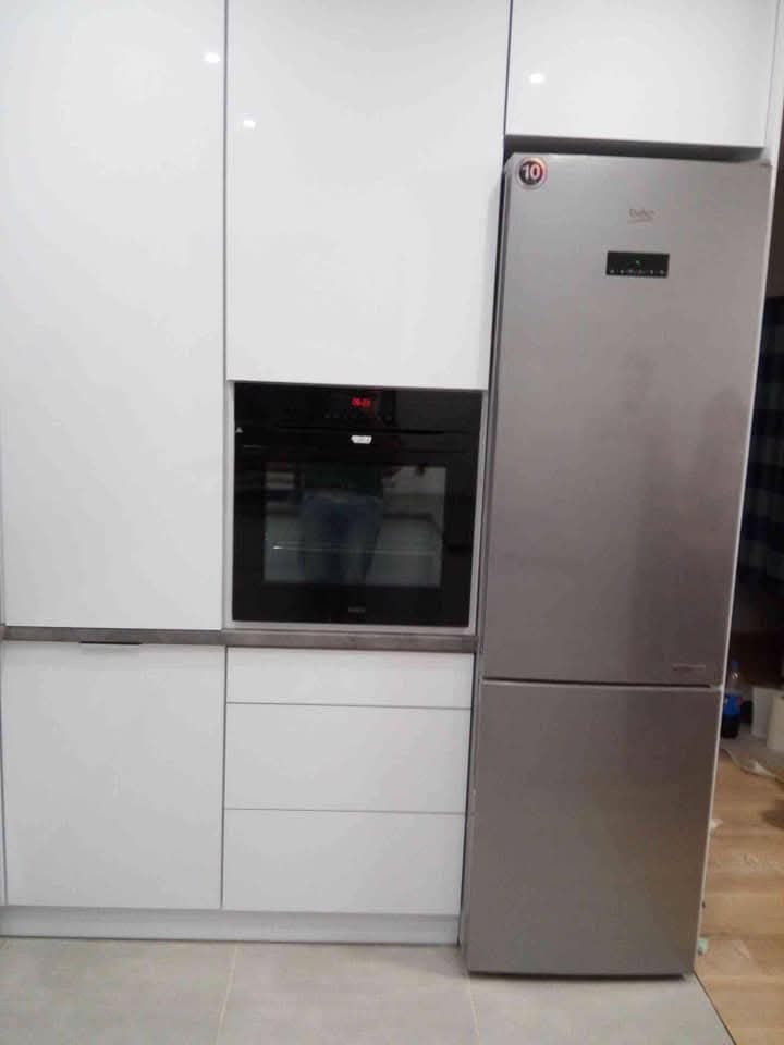 Biała zabudowa kuchenna z piekarnikiem i lodówką Beko. Minimalistyczny design, gładkie fronty, szare blaty. Ustawienie sprzętów AGD w zabudowie.