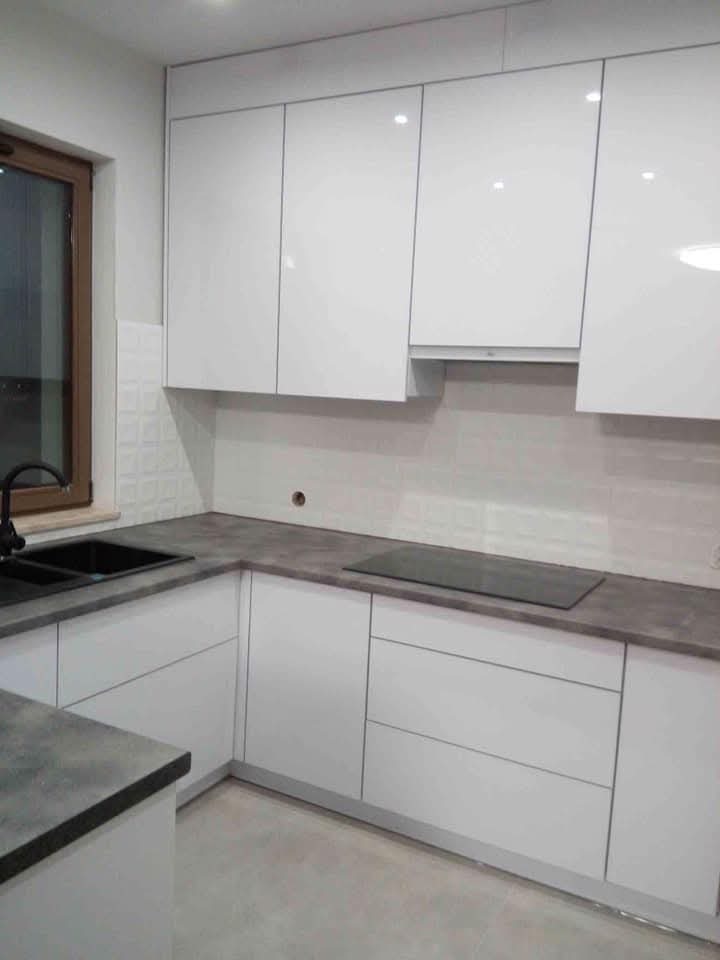 Biała, minimalistyczna kuchnia z ciemnym blatem i czarnym zlewem, widziana pod kątem. Górne szafki lakierowane na wysoki połysk, brak uchwytów. Okno w tle.