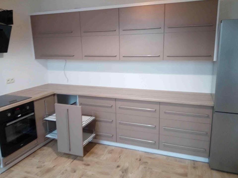 Beżowa kuchnia z wysuwaną szafką narożną i srebrnymi uchwytami, widok na górne i dolne szafki, drewniana podłoga, nowoczesny design.