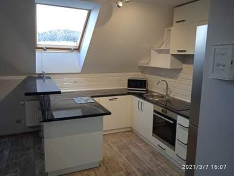 Biała kuchnia na poddaszu ze skośnym oknem dachowym, czarnym blatem i drewnianą podłogą. Zabudowa kuchenna dopasowana do skosów.