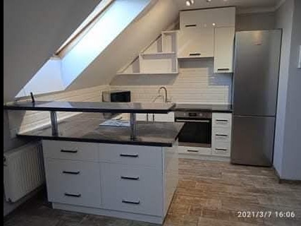 Funkcjonalna kuchnia na poddaszu ze skośnym oknem dachowym. Białe szafki, czarny blat, nowoczesna wyspa i srebrna lodówka tworzą spójną całość.