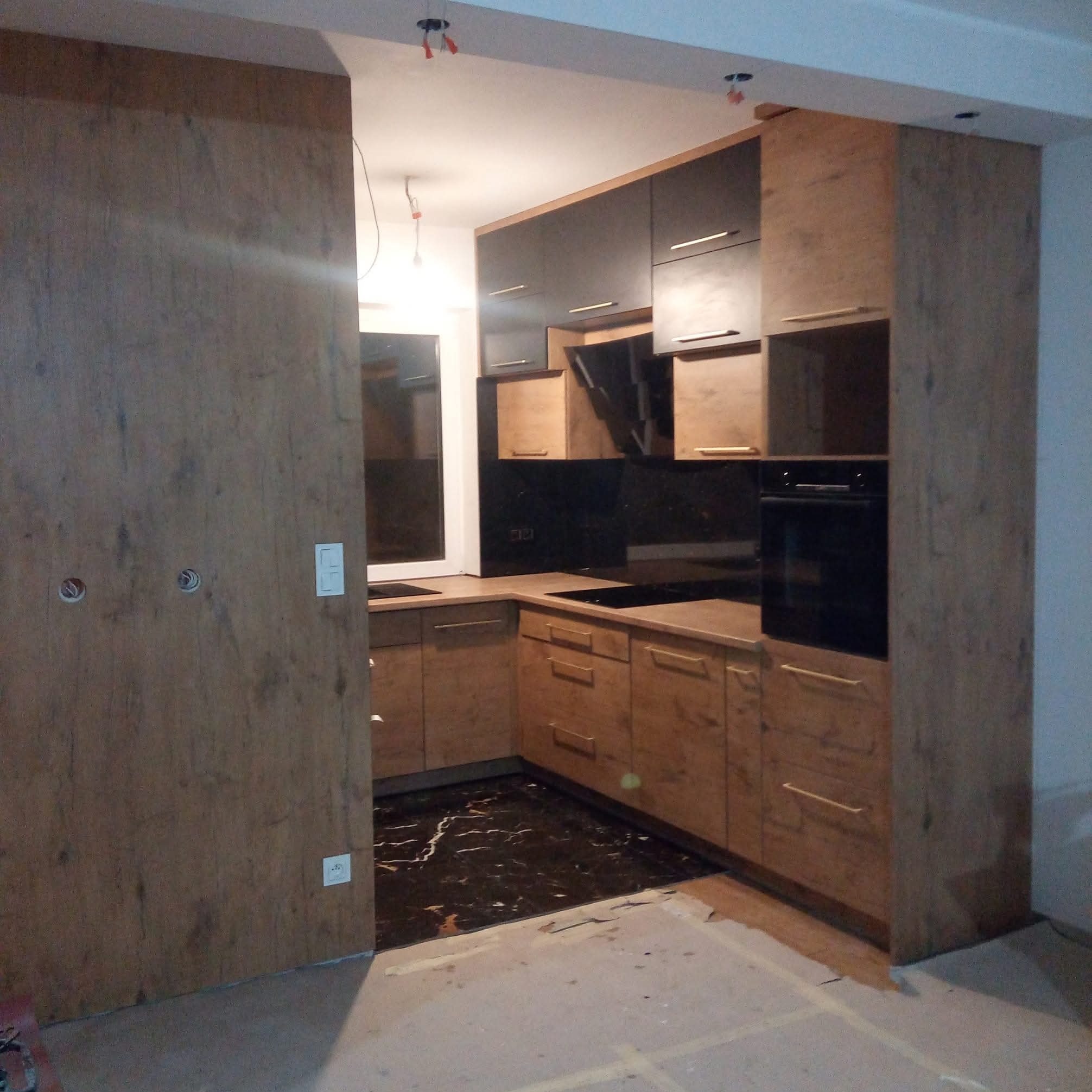 Nowoczesna kuchnia z forniru w odcieniach brązu i czerni, z czarnym blatem i marmurową podłogą. Widoczne przygotowane punkty oświetleniowe na suficie.