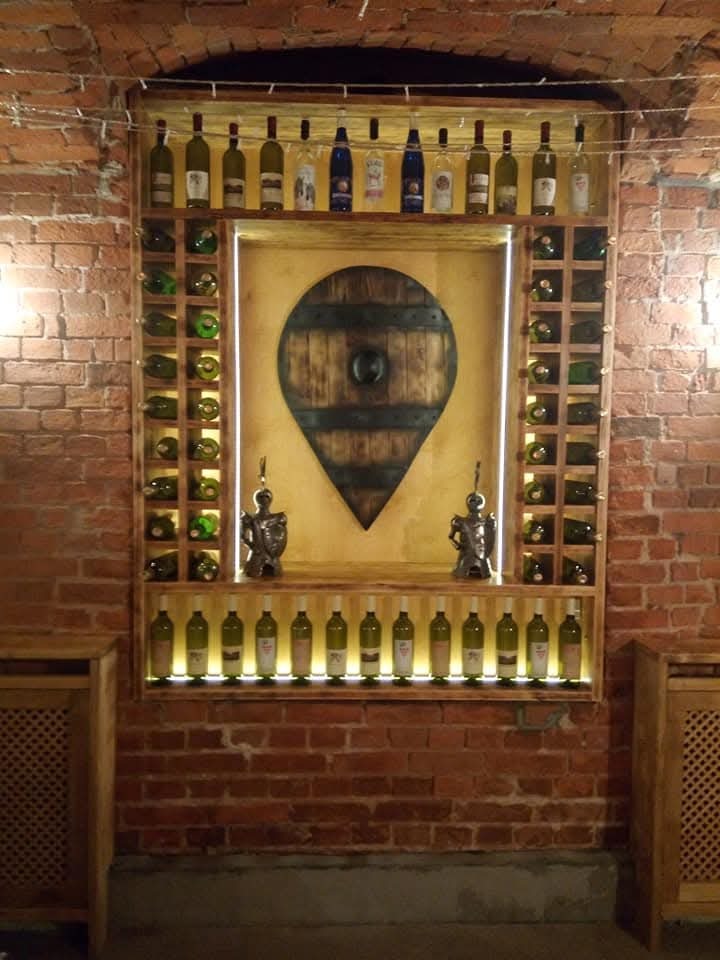 Ręcznie robiona, podświetlana półka na wino w stylu rustykalnym, z drewnianym panelem dekoracyjnym w kształcie łzy, umieszczona na tle ściany z czerwonej cegły.