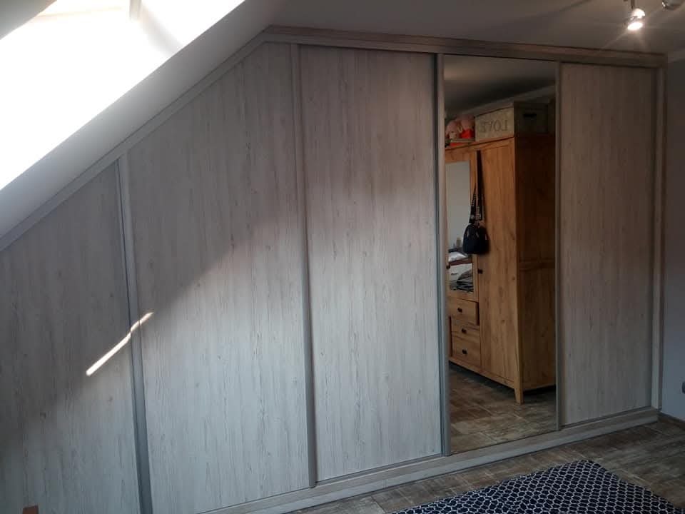 Jasna szafa wnękowa z przesuwnymi drzwiami i lustrem, dopasowana do skosu dachu. Widoczny fragment podłogi i oświetlenie sufitowe.