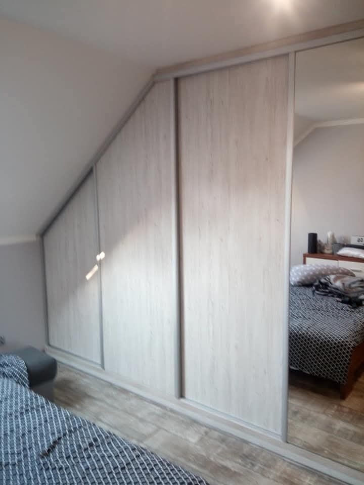 Biała szafa wnękowa z przesuwnymi drzwiami, dopasowana do skosu poddasza, z jednym lustrzanym skrzydłem, odbijającym fragment sypialni.