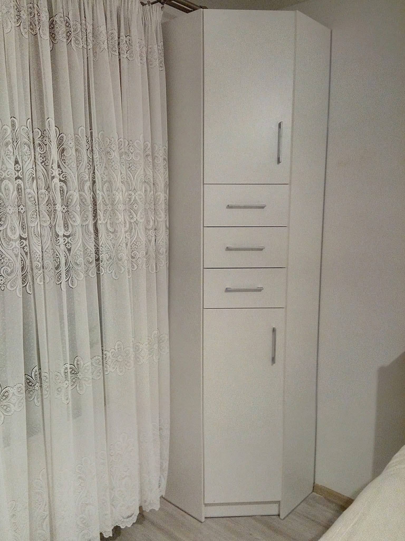 Biała szafa narożna z szufladami i drzwiczkami, widoczna obok delikatnej, wzorzystej firanki. Minimalistyczny design, jasne wnętrze, subtelne uchwyty.