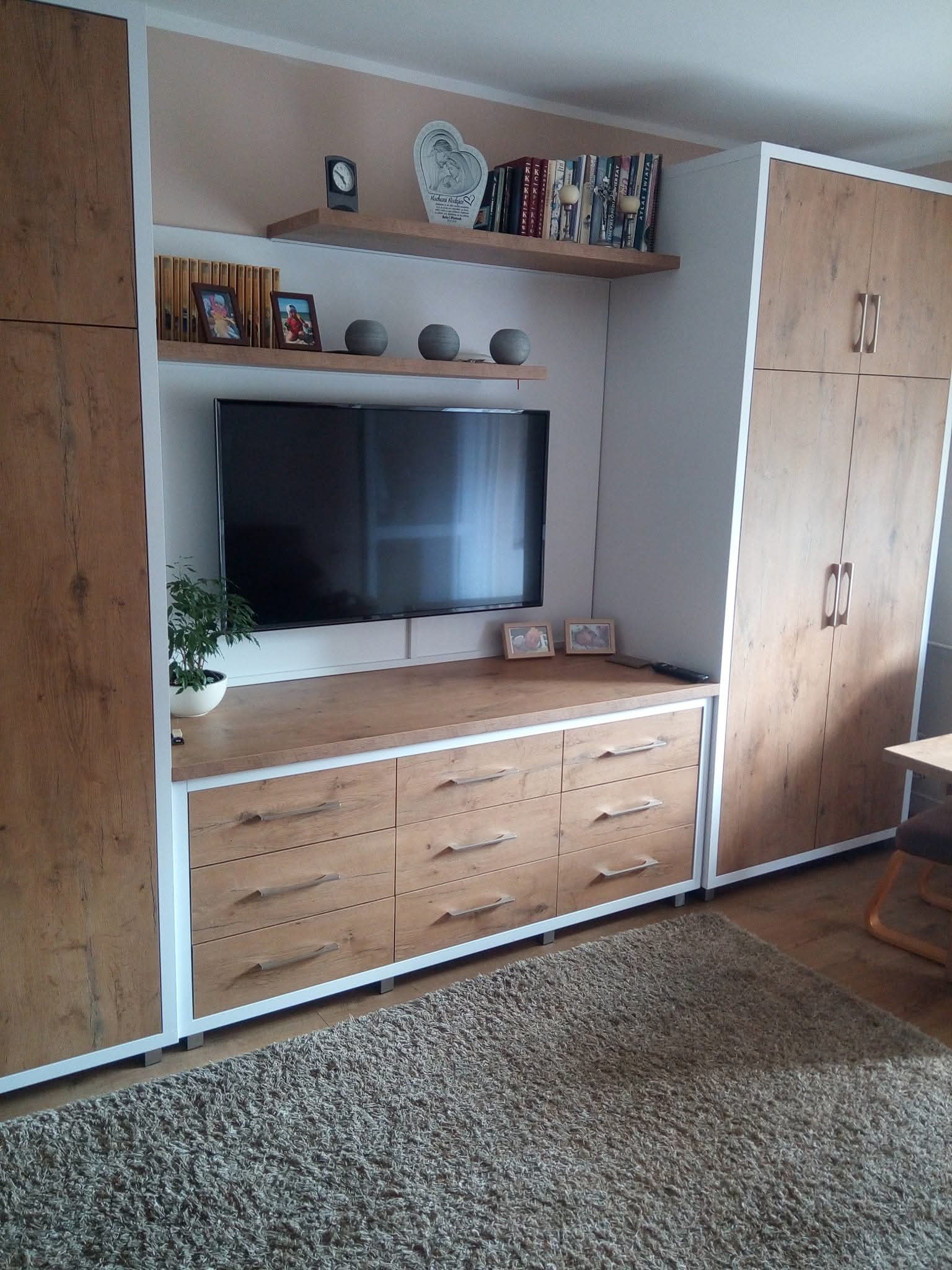 Zabudowa meblowa w salonie z szafą, komodą z szufladami i półkami, utrzymana w odcieniach drewna z białymi akcentami. Na półkach dekoracje i zdjęcia.