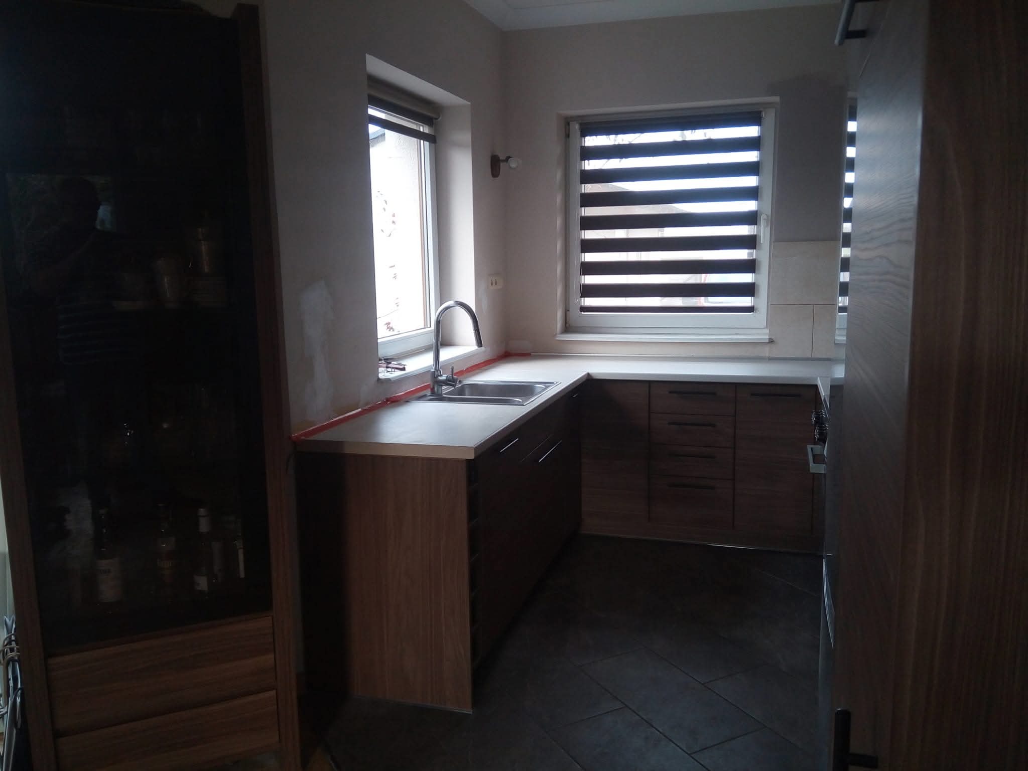 Kuchnia w odcieniach brązu z drewnianymi frontami, widok na zlew pod oknem z roletami, ciemna podłoga z płytek imitujących drewno, oświetlenie naturalne.