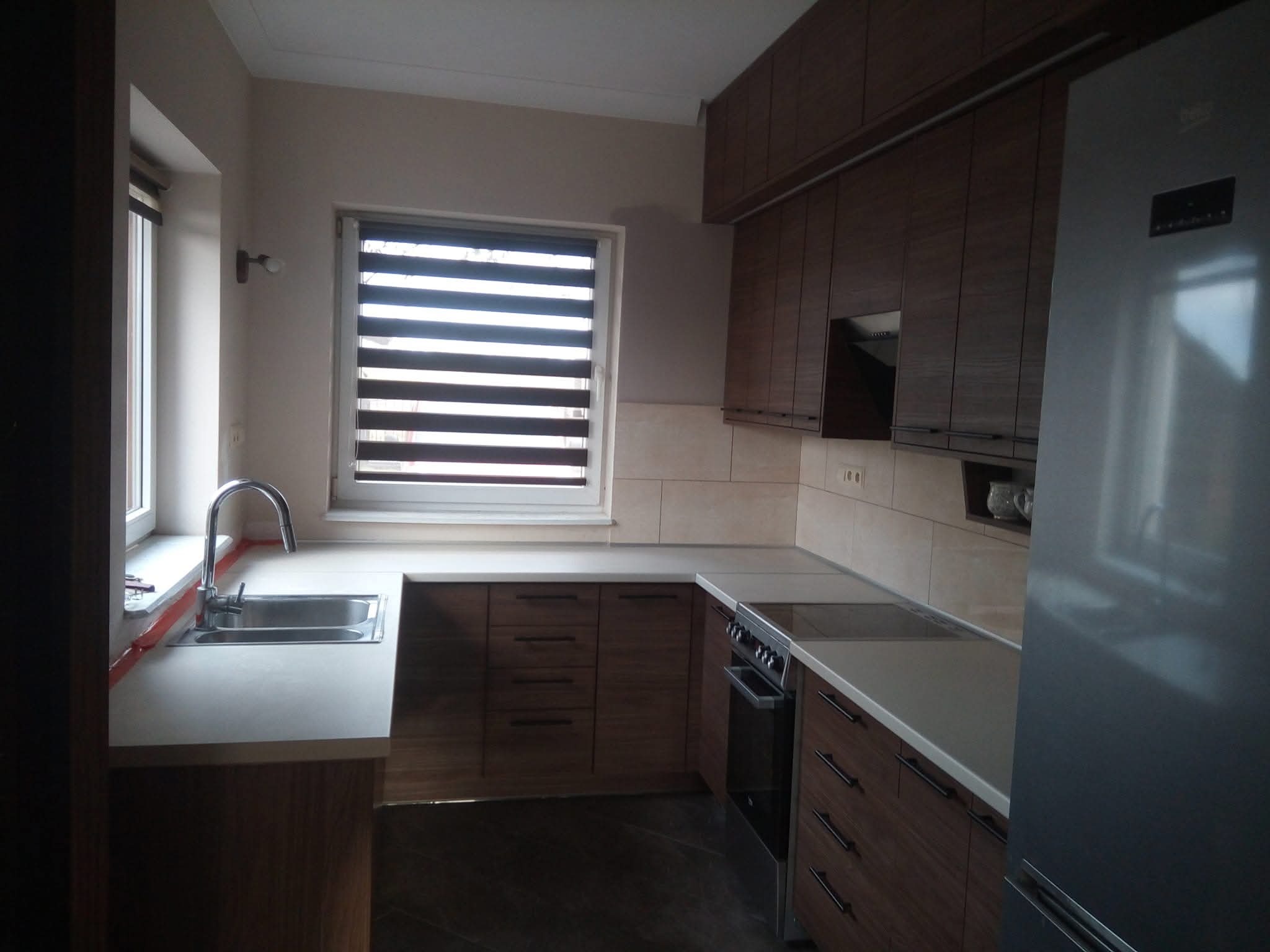 Nowoczesna kuchnia z drewnianymi szafkami, jasnym blatem i roletami w oknie. Widoczny zlew, płyta grzewcza i lodówka. Uwagę zwraca spójny design i funkcjonalność.