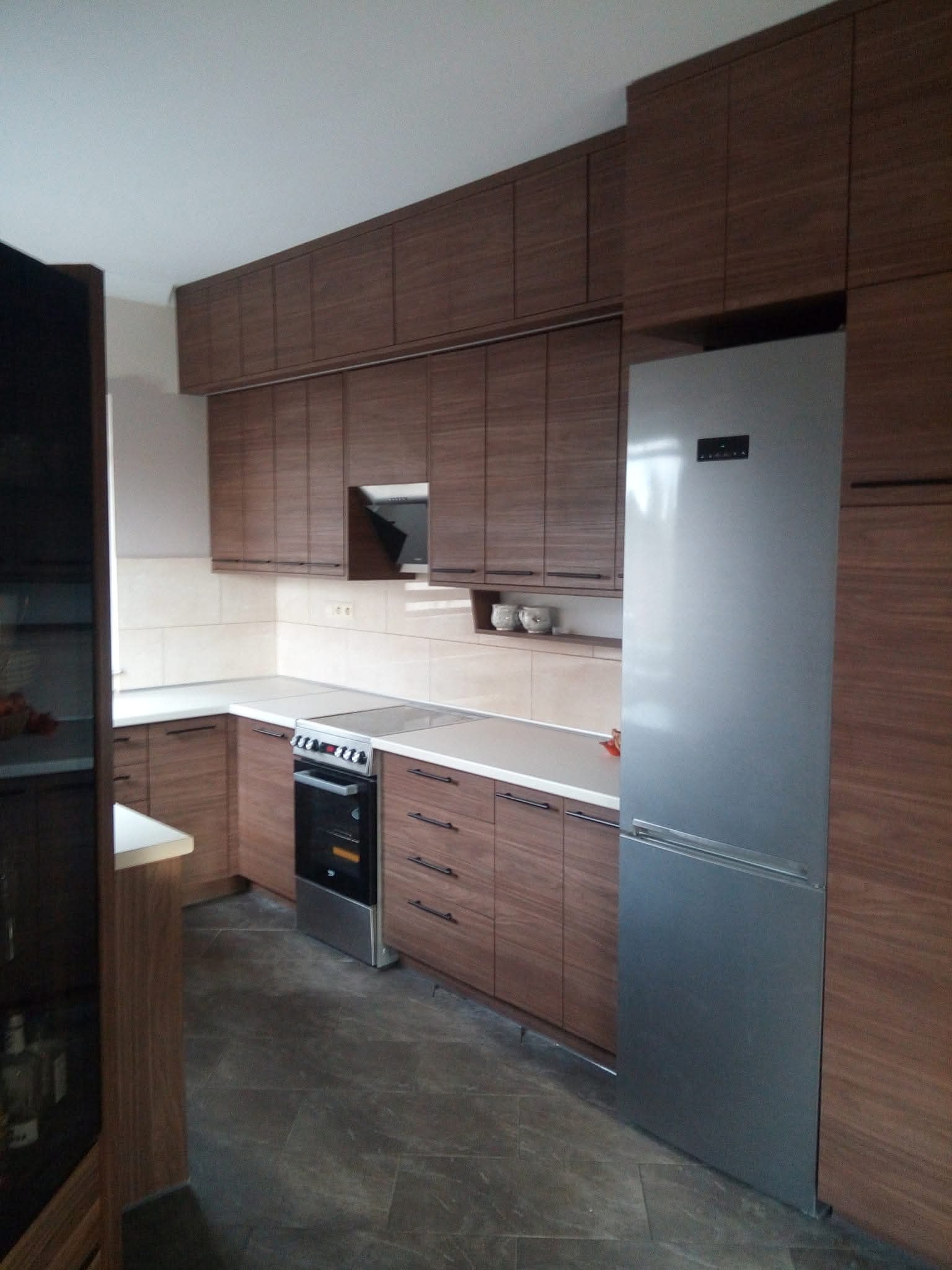 Nowoczesna kuchnia w odcieniach brązu z drewnianymi frontami i srebrną lodówką. Górne szafki wiszące, dolne z szufladami. Płytki ceramiczne na ścianie.