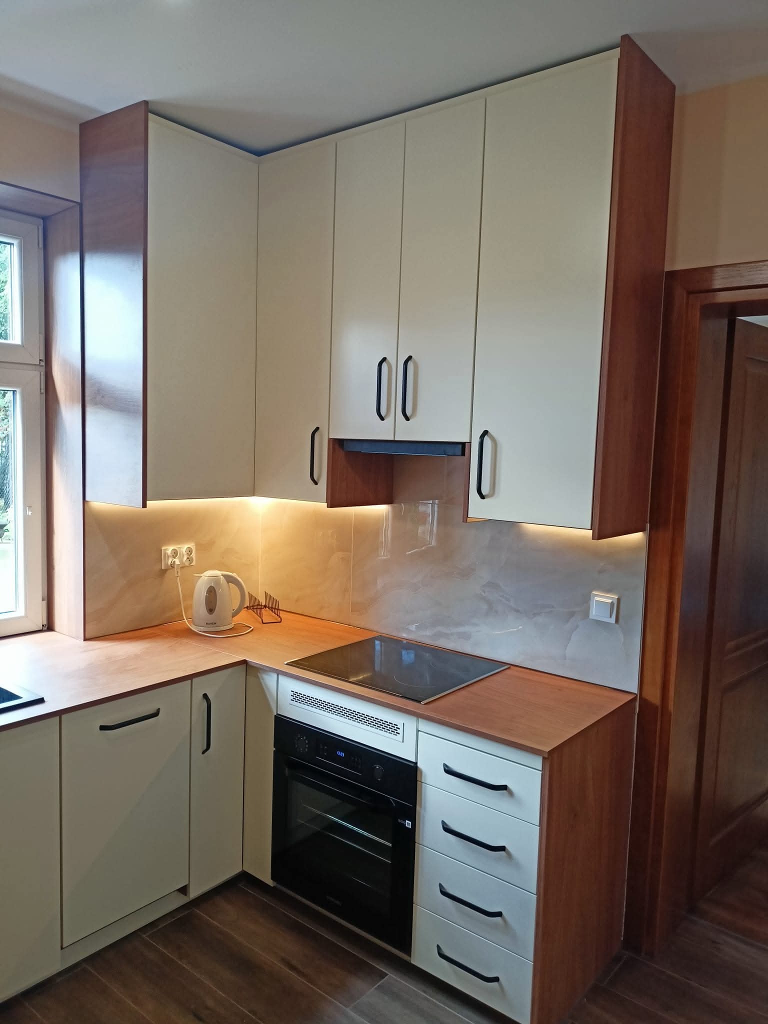 Nowoczesna kuchnia narożna z drewnianymi akcentami, oświetleniem LED podszafkowym i sprzętem AGD w zabudowie. Jasne fronty szafek i ciemne, podłużne uchwyty.