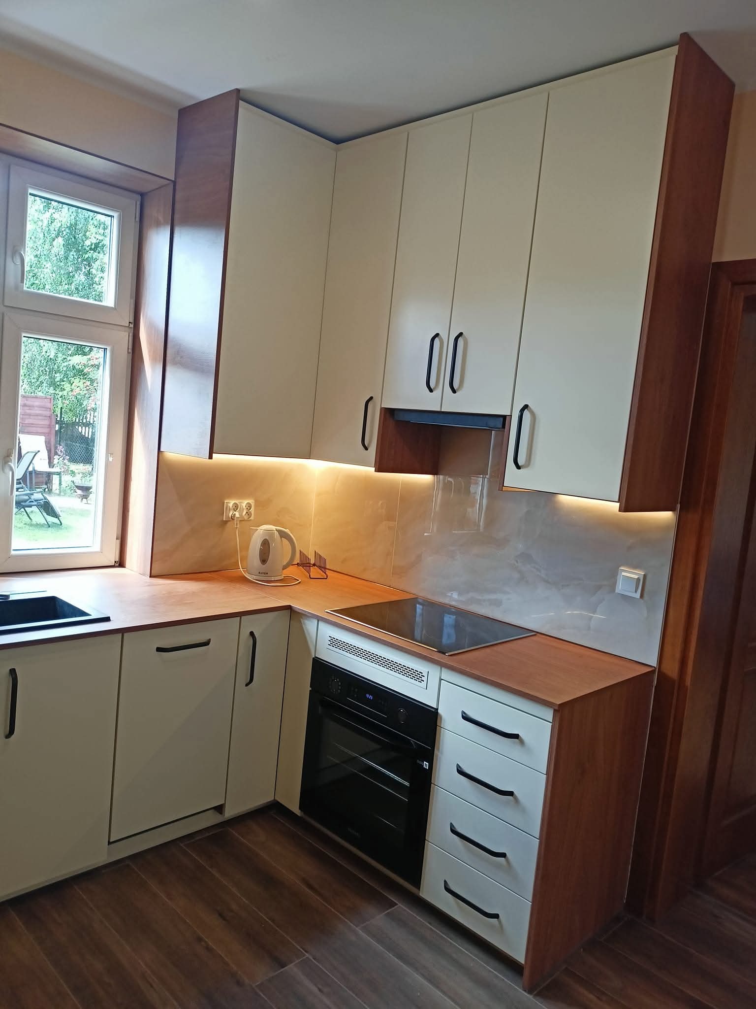 Jasna kuchnia z drewnianymi akcentami: zabudowa meblowa z oświetleniem LED podszafkowym, płyta indukcyjna i okno z widokiem na ogród. Nowoczesny minimalizm.