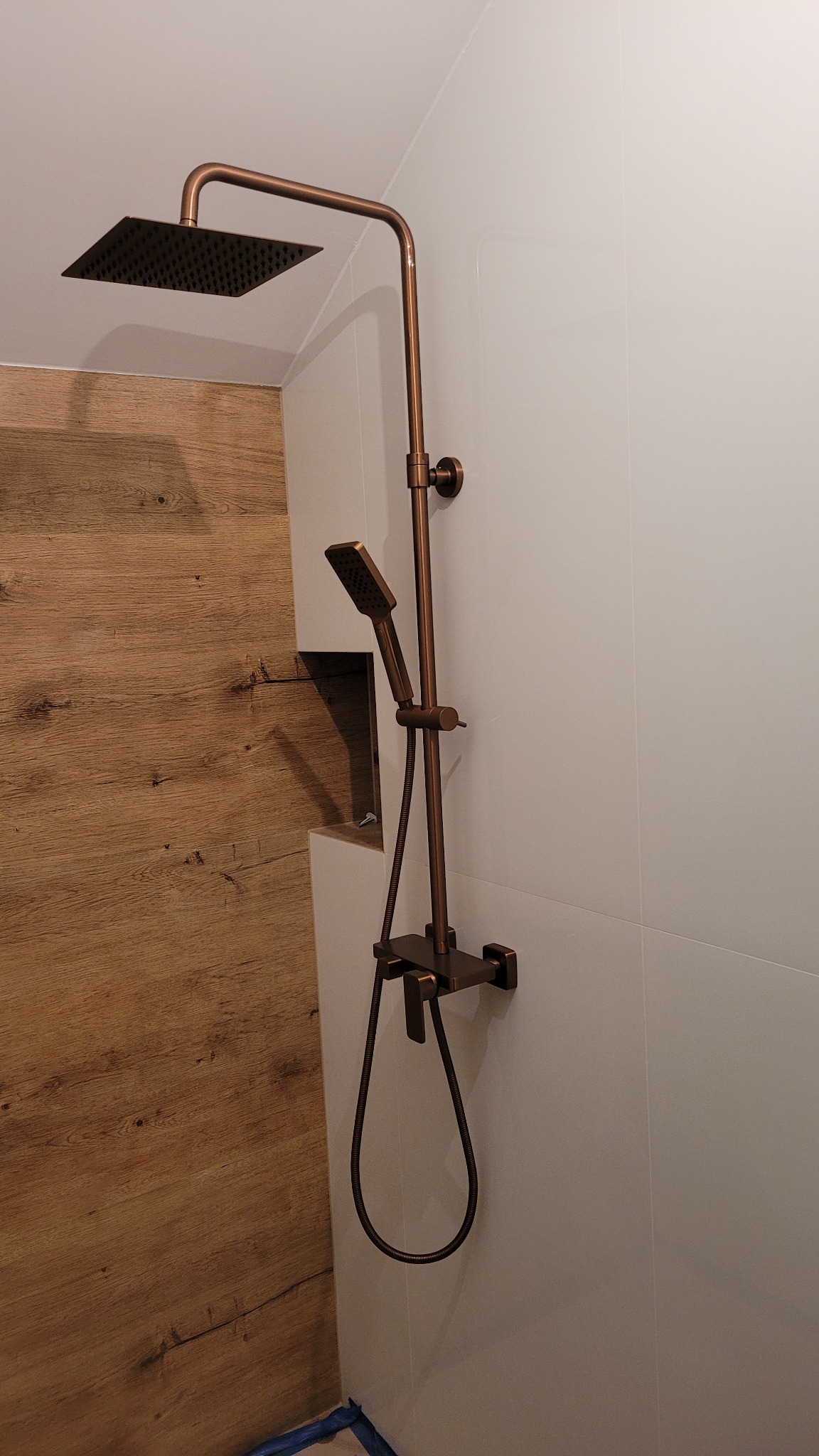 Miedziana bateria prysznicowa z deszczownicą i słuchawką, zamontowana na białej ścianie z płytek, obok drewnianej wnęki.