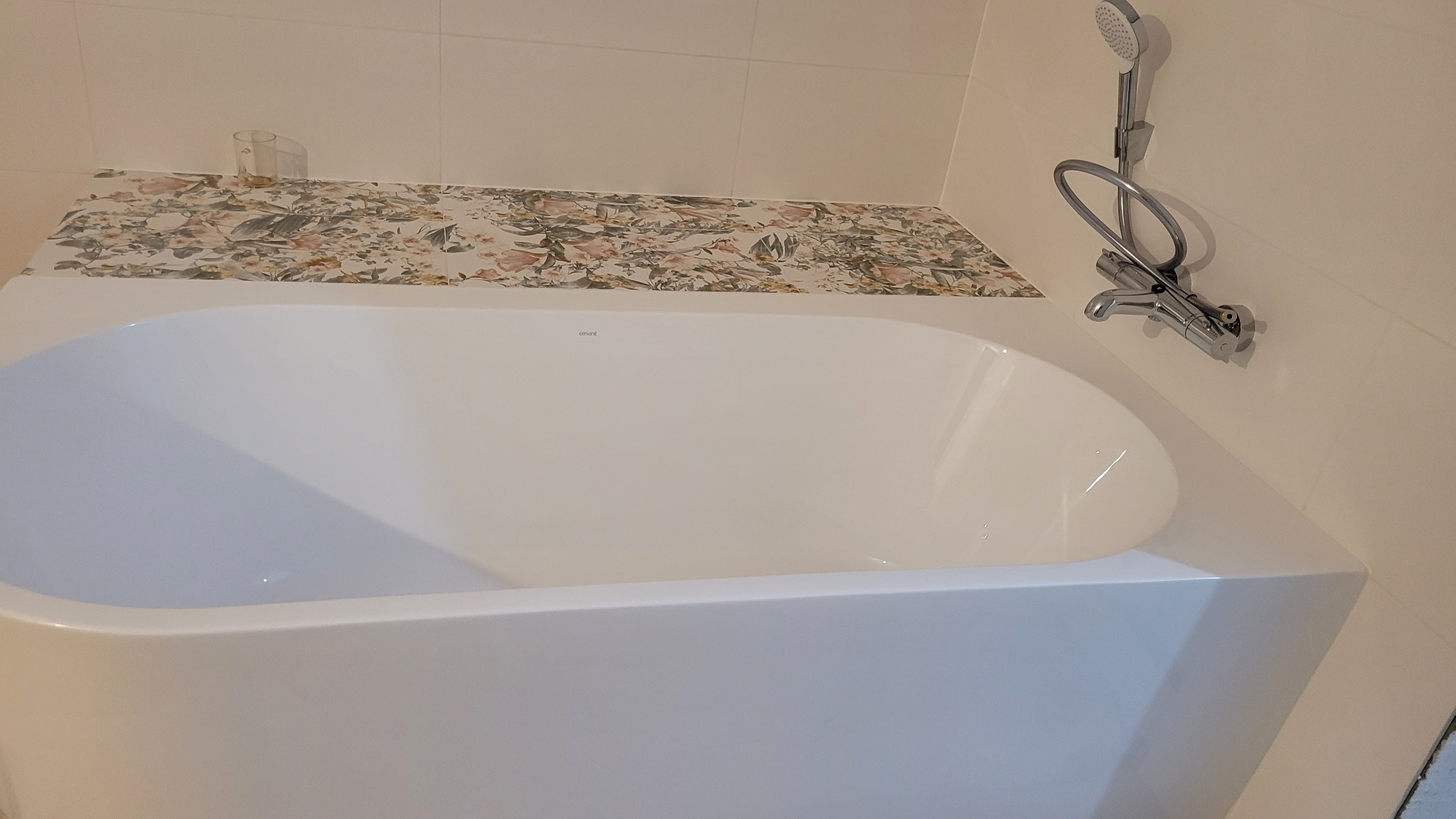 Biała wanna z armaturą i słuchawką prysznicową na tle płytek z motywem kwiatowym. Widok z góry prezentuje nową instalację w łazience.