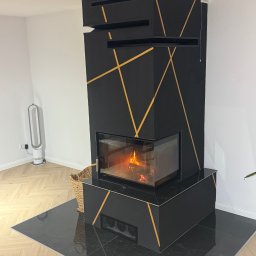 BACA BACA - Czarny kominek z geometrycznym, złotym zdobieniem w Ząb. Nowoczesny design, widoczny płomień. Podłoga wyłożona czarnym gresem.