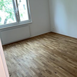 BACA BACA - Jasne, drewniane panele podłogowe w pokoju z białymi ścianami i grzejnikiem pod oknem. Wisząca żarówka dodaje surowego charakteru wnętrzu.