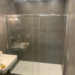 Nowoczesna kabina prysznicowa z przesuwnymi drzwiami, wykończona szarą płytką, z widoczną armaturą i umywalką. Minimalistyczny design, oświetlenie punktowe.