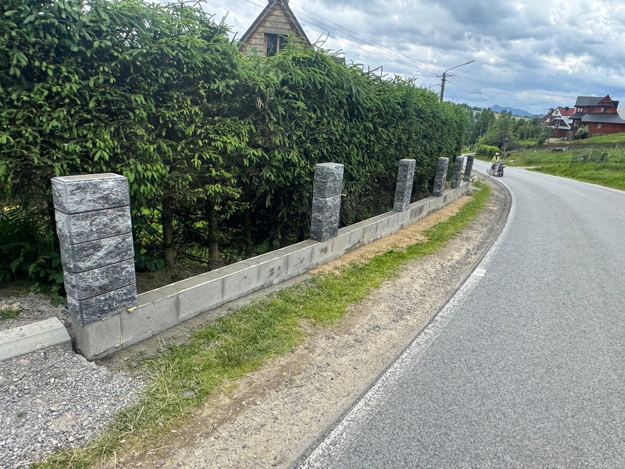 Nowo budowany, niski murek z betonowych elementów i kamiennych słupków, oddzielający posesję od drogi. W tle żywopłot i domy w górskim krajobrazie.