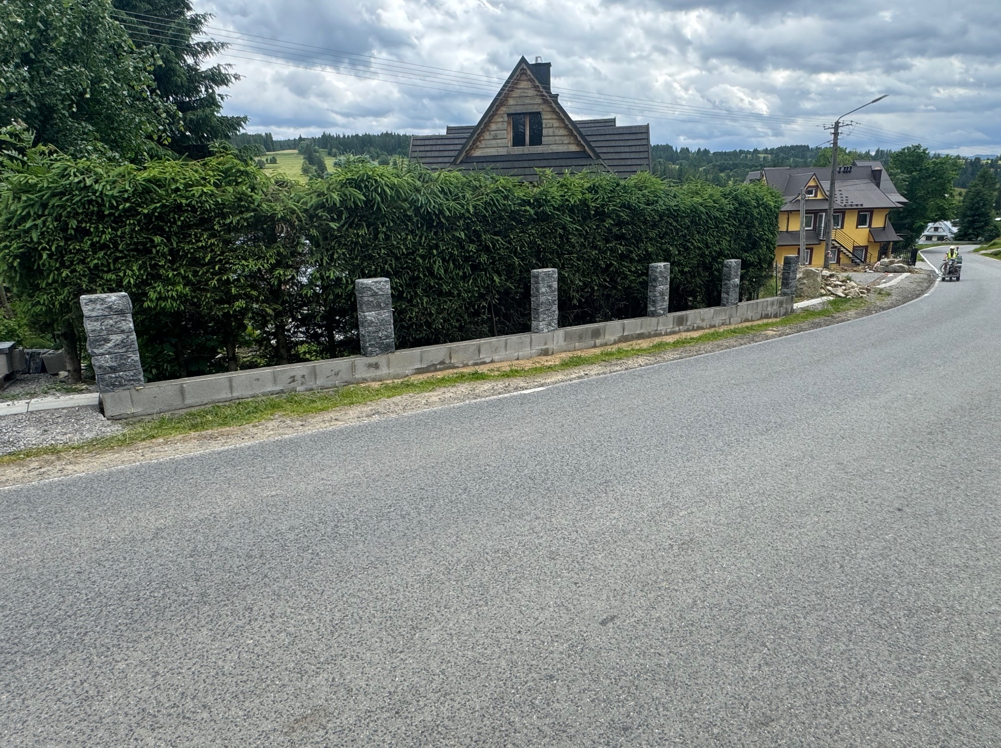 Nowo wybudowany, niski murek z szarej kostki z nasadzeniami z krzewów, oddzielający posesję od drogi. W tle domy w górskim stylu i pochmurne niebo.