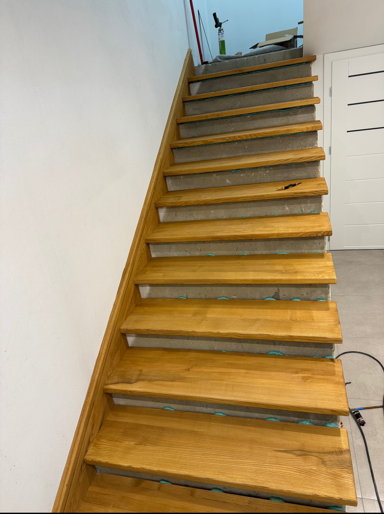 Montaż drewnianych schodów na betonowej konstrukcji. Widoczne pianowanie szczelin, biała ściana i drzwi w tle. Ujęcie z dołu, schody w trakcie prac wykończeniowych.