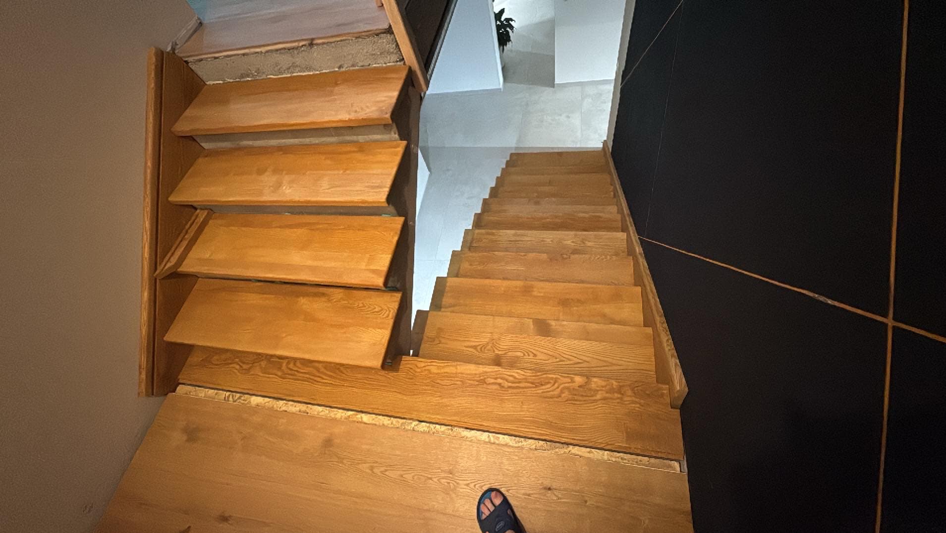 Drewniane schody z widoczną strukturą drewna, prowadzące na wyższą kondygnację. Obok ściana w kolorze czarnym z ozdobnymi, złotymi liniami.