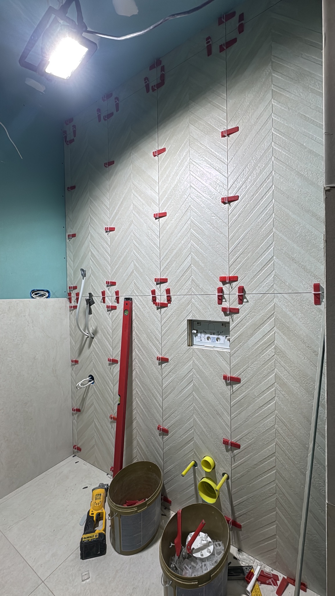 Układanie płytek w jodełkę w łazience, z użyciem poziomicy i klipsów dystansowych. Widoczne narzędzia i wiaderka, sufit pomalowany na niebiesko.